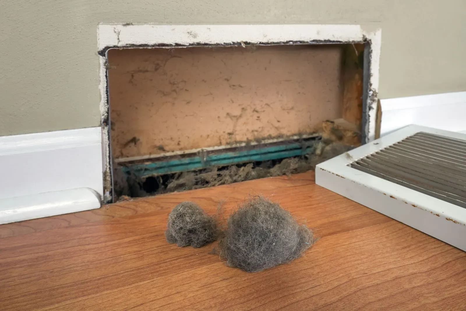 Dust bunnies on hardwood floor in front of open wall vent, illustrating need for home maintenance.
