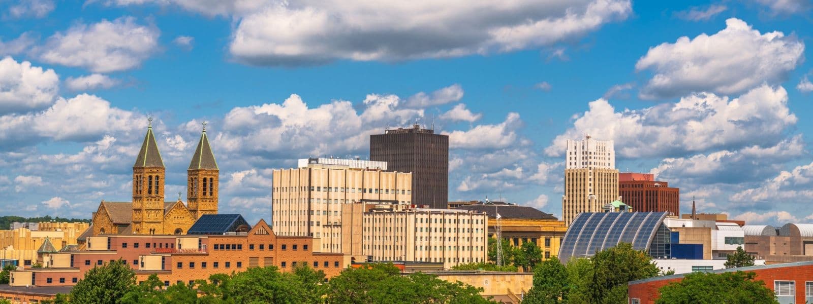 Skyline of Akron, OH showing a church with twin spires on the left, mid-rise office and residential buildings in the center, and modern structures on the right, under a bright blue sky with white clouds.