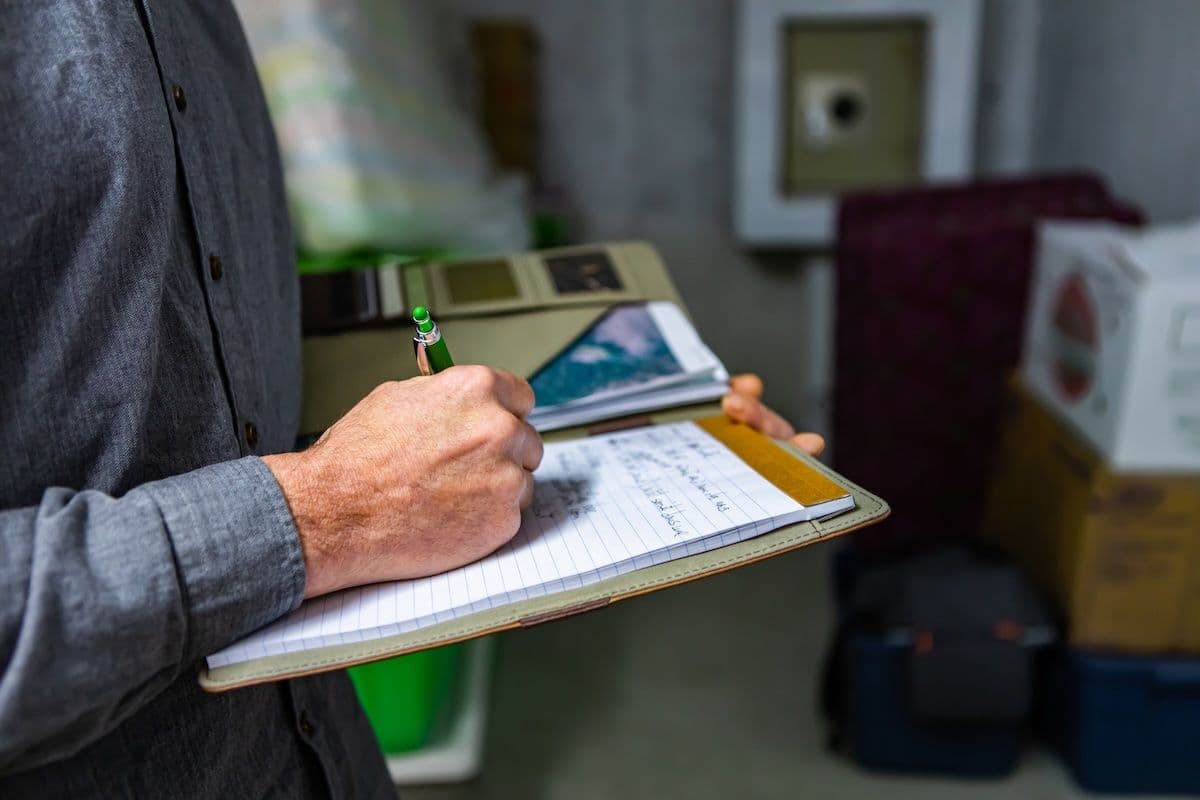 Person holding clipboard with pen, writing on lined paper in an office or institutional setting.