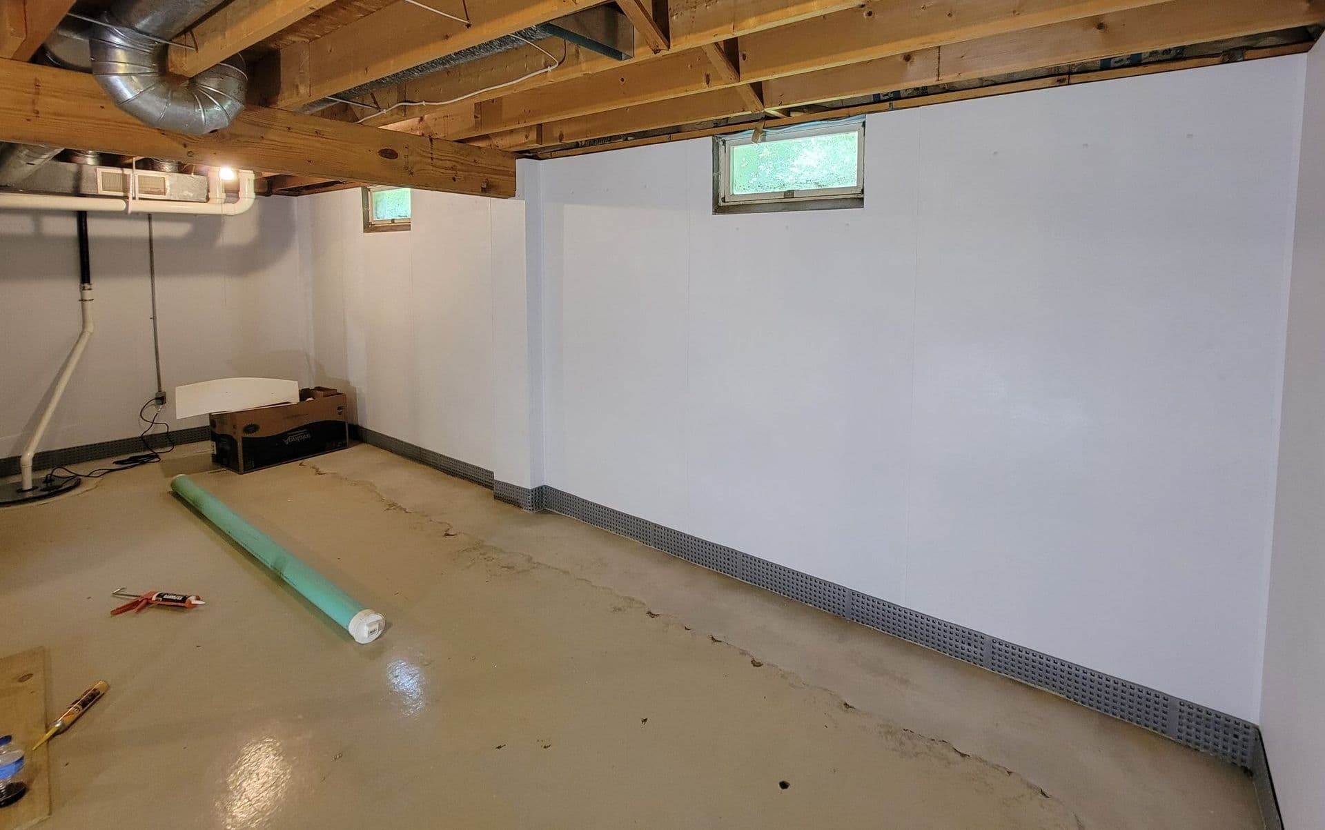 Unfinished basement under renovation with concrete floor, white drywall, exposed wooden beams, small windows, PVC pipe, and metal baseboard trim.