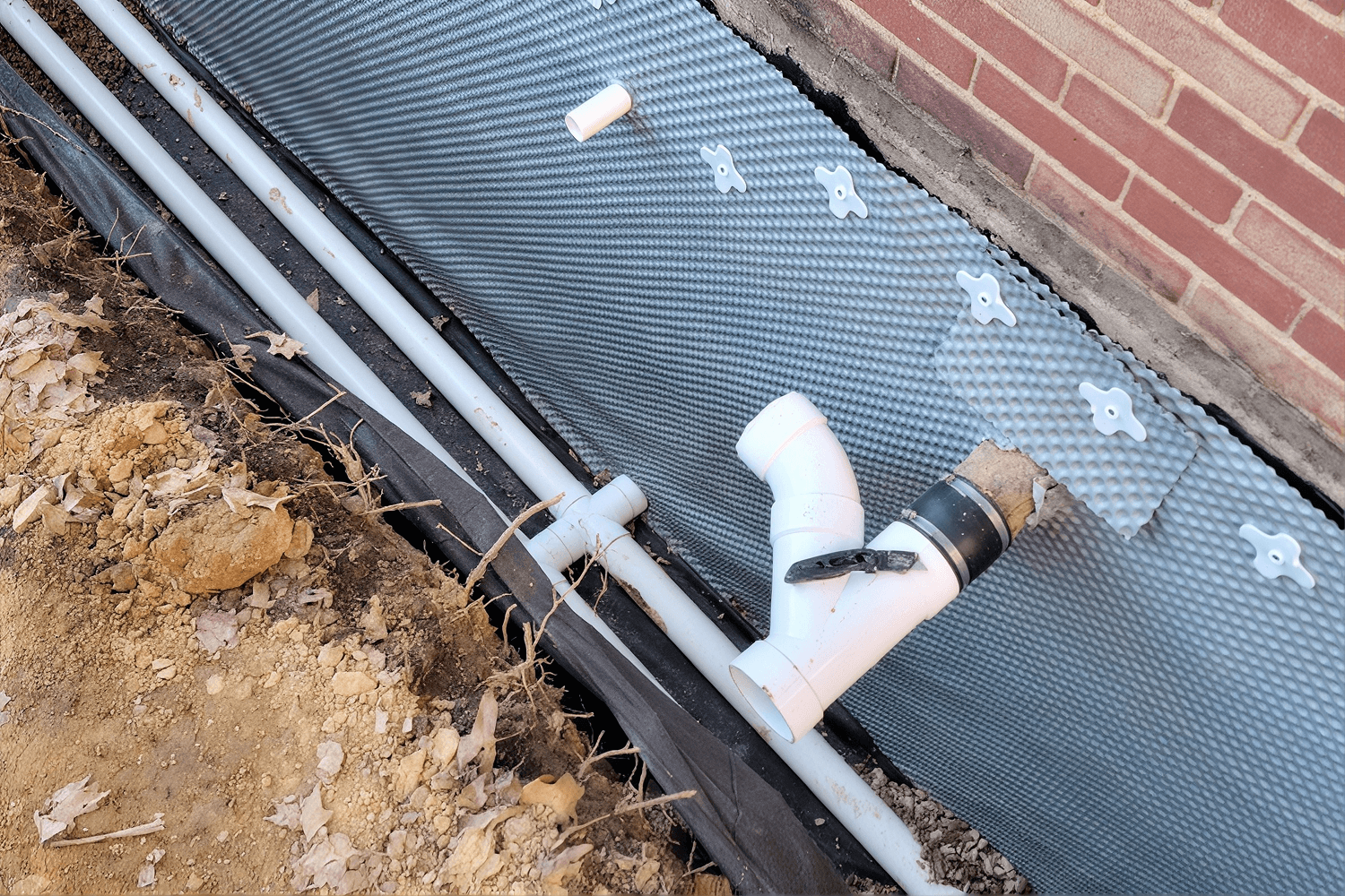 Excavated foundation with white PVC drainage pipes and elbow connector installed next to brick foundation and soil.