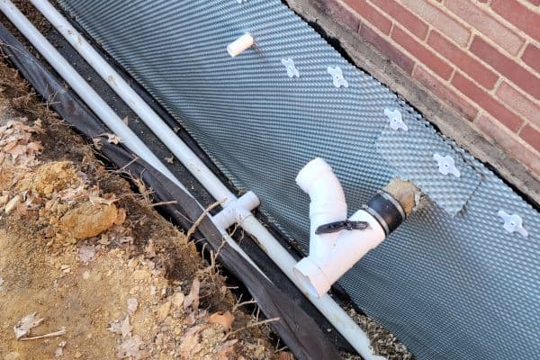 Basement window well with white PVC drainage pipe, protective mesh covering, and bare soil alongside red brick foundation.