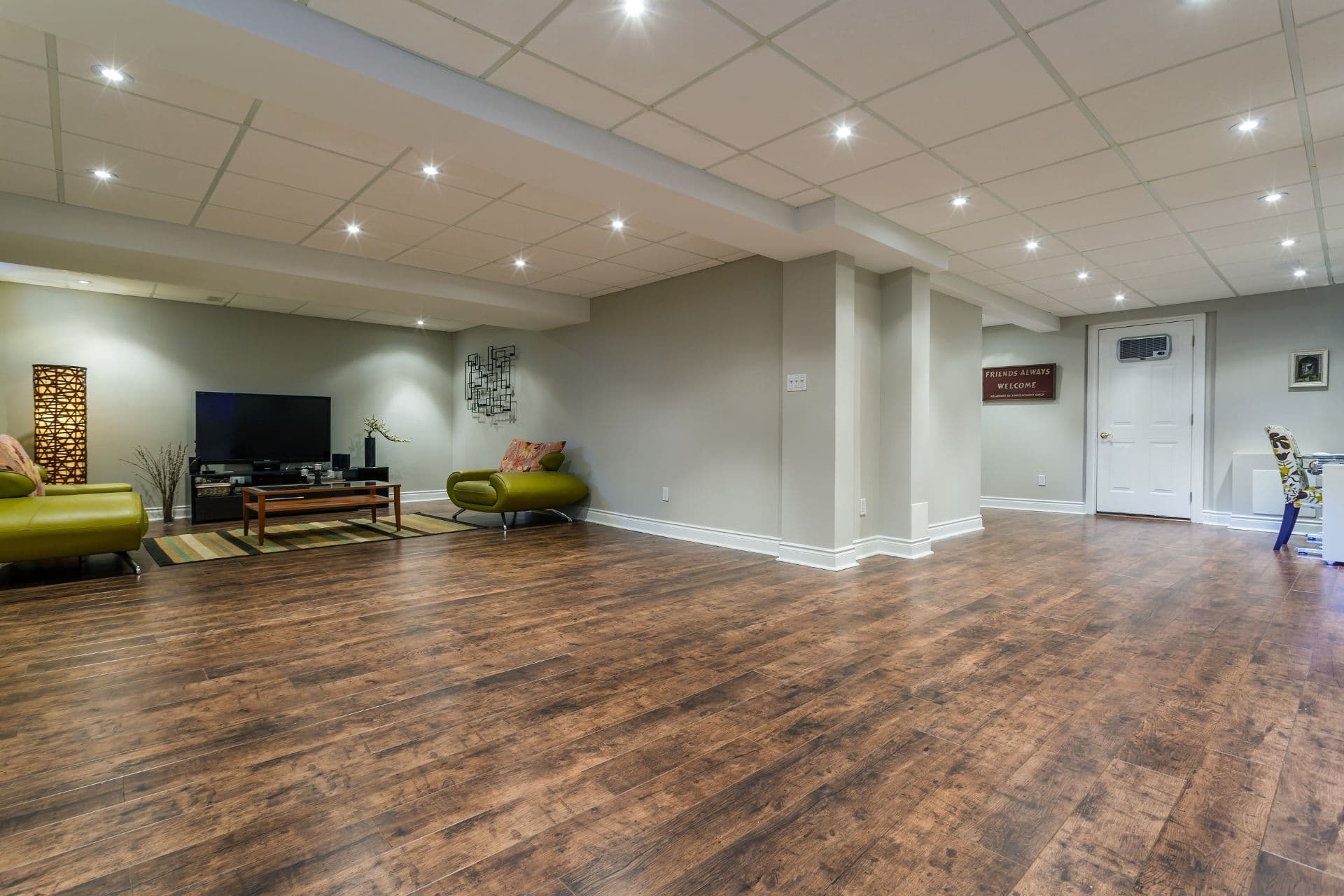 Spacious finished basement family room with wood-look flooring, recessed ceiling lights, green upholstered seating, TV entertainment center, white support columns, and doorways leading to other areas.