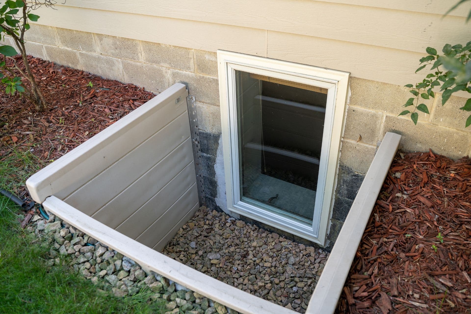 Basement egress window with white frame installed in concrete foundation wall, surrounded by gravel and mulch landscaping.