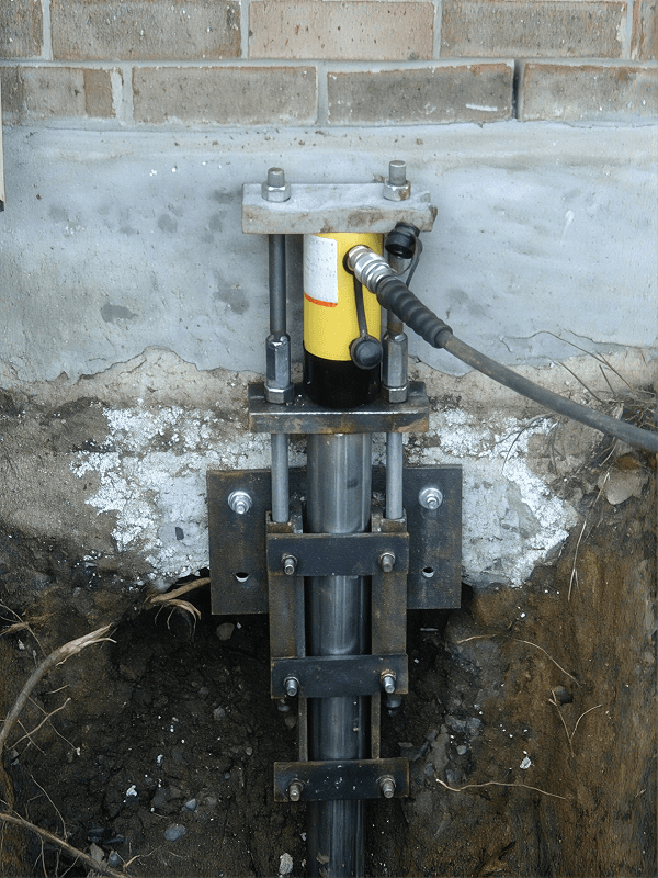 Yellow and black hydraulic jack mounted on steel post against concrete foundation wall with attached hose and bolted mounting hardware.