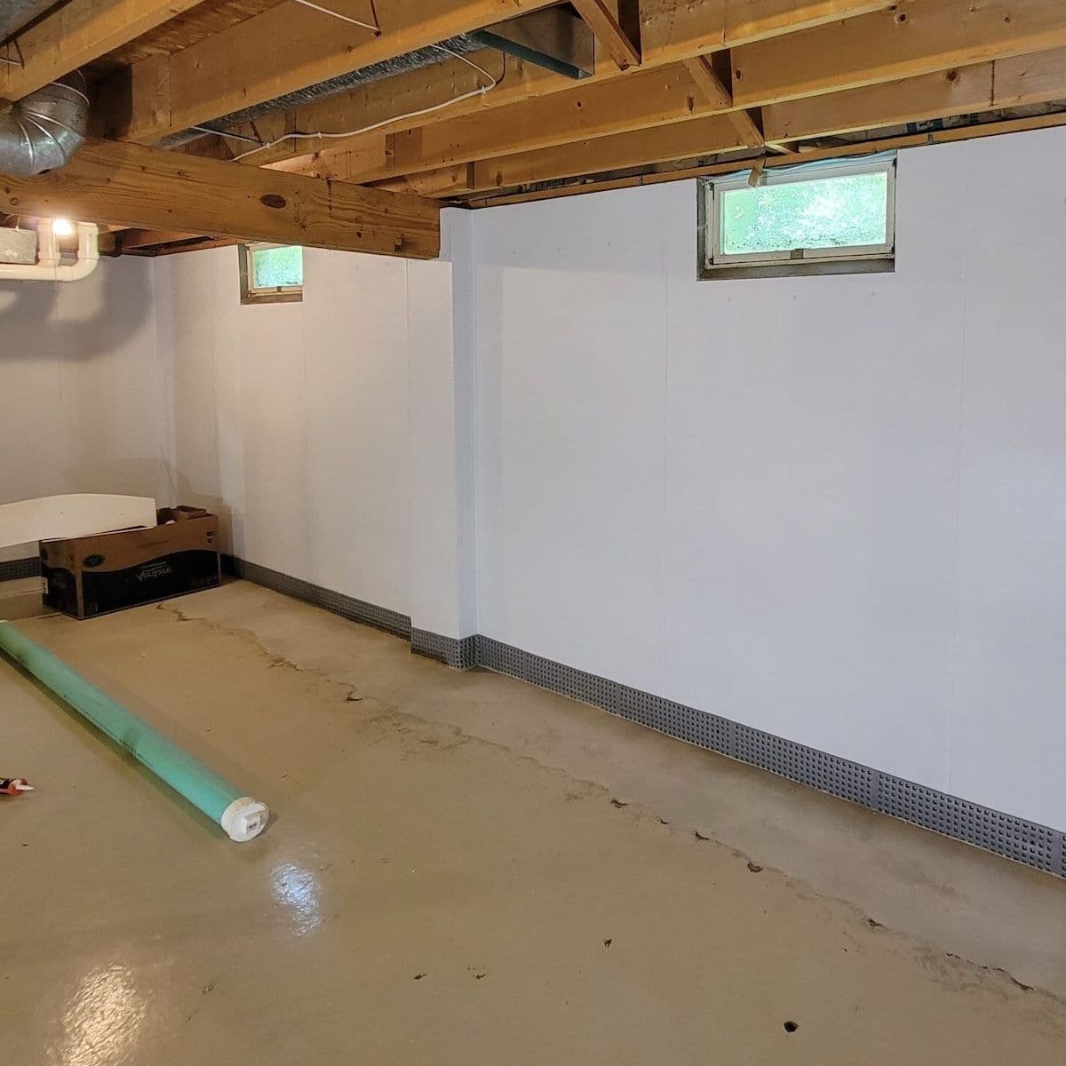 Unfinished basement with white drywall-lined walls, concrete floor, exposed wooden beams, small windows, and protective floor trim along the baseboard.
