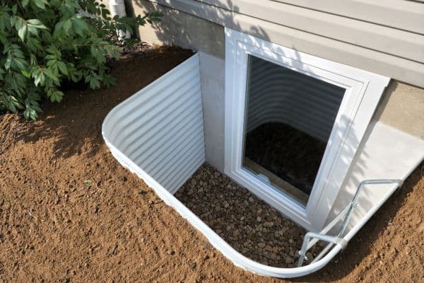 White metal basement egress window well with open cover and dark opening leading underground, set in brown soil next to house foundation.