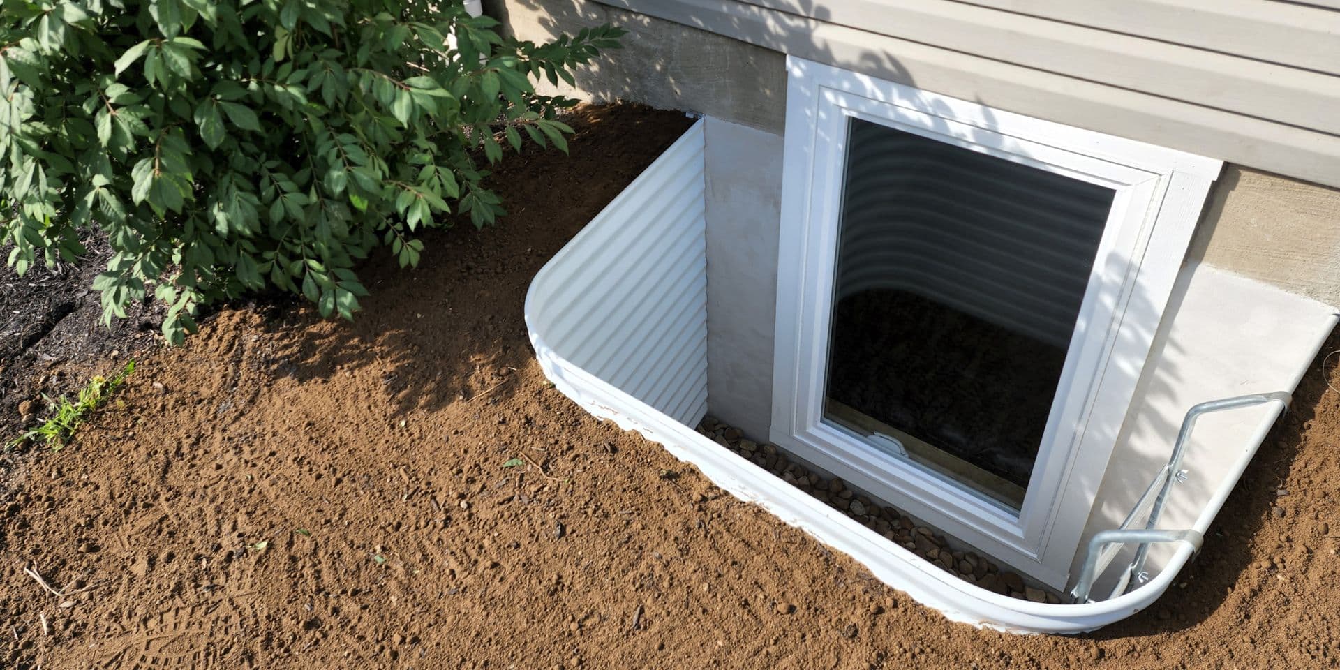 White metal basement egress window well attached to house foundation, surrounded by brown soil and green foliage.