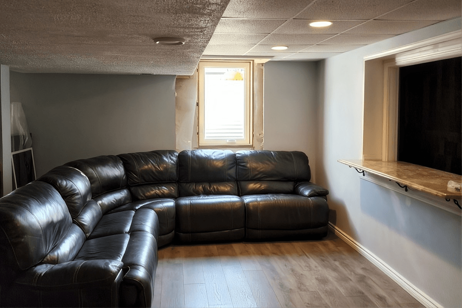 Finished basement recreation room with black leather sectional sofa, wooden flooring, recessed ceiling lights, and egress window with natural light.