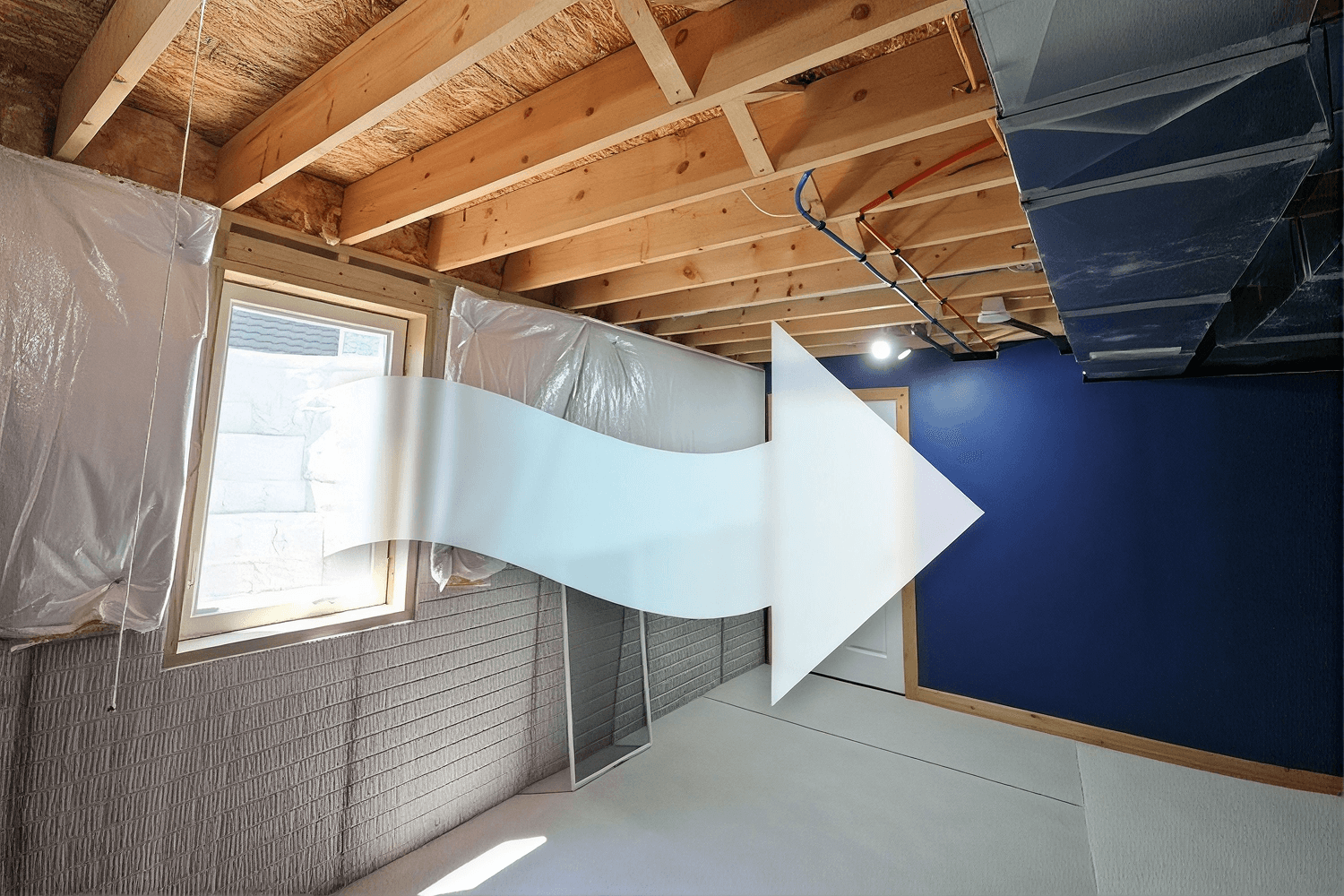 Unfinished basement with exposed wood ceiling joists, white plastic sheeting, blue painted wall, and large white arrow pointing right from an egress window.