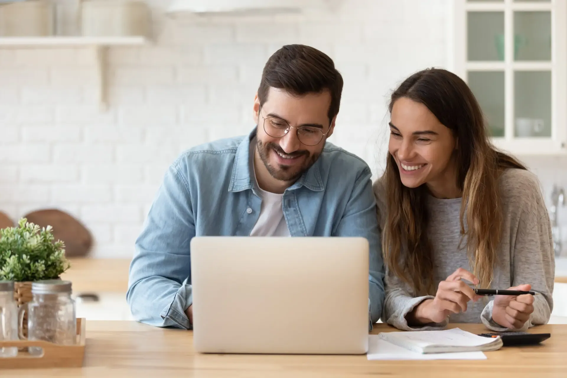 Couple smiling while reviewing content on laptop at wooden desk in bright kitchen setting.