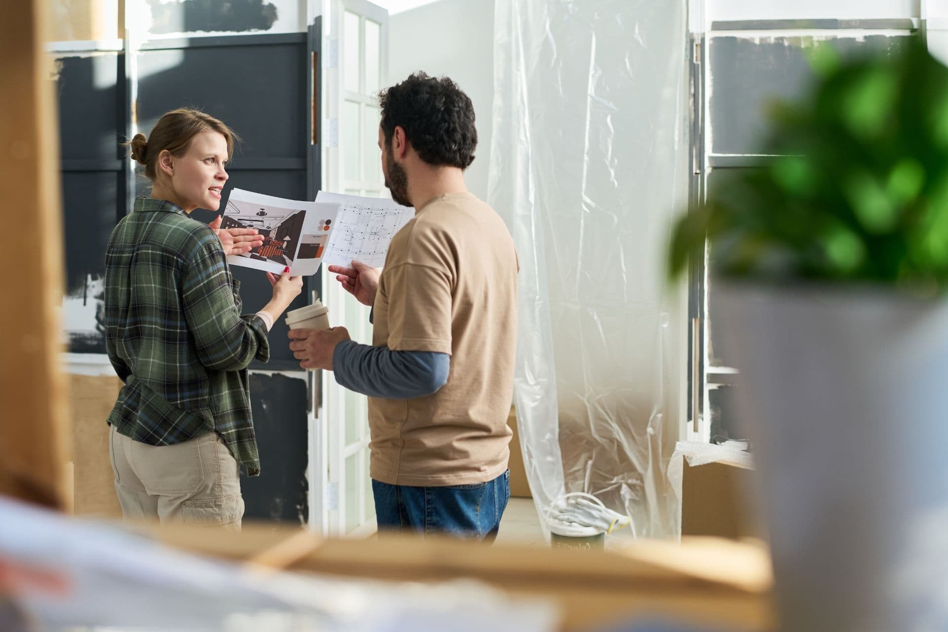 Two people review construction plans and blueprints together on a renovation jobsite with plastic sheeting and windows visible in background.