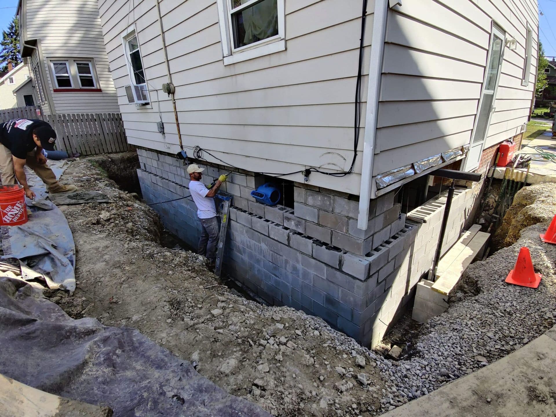 Workers installing foundation repair system on house exterior with excavated trench and cinder block wall reinforcement.