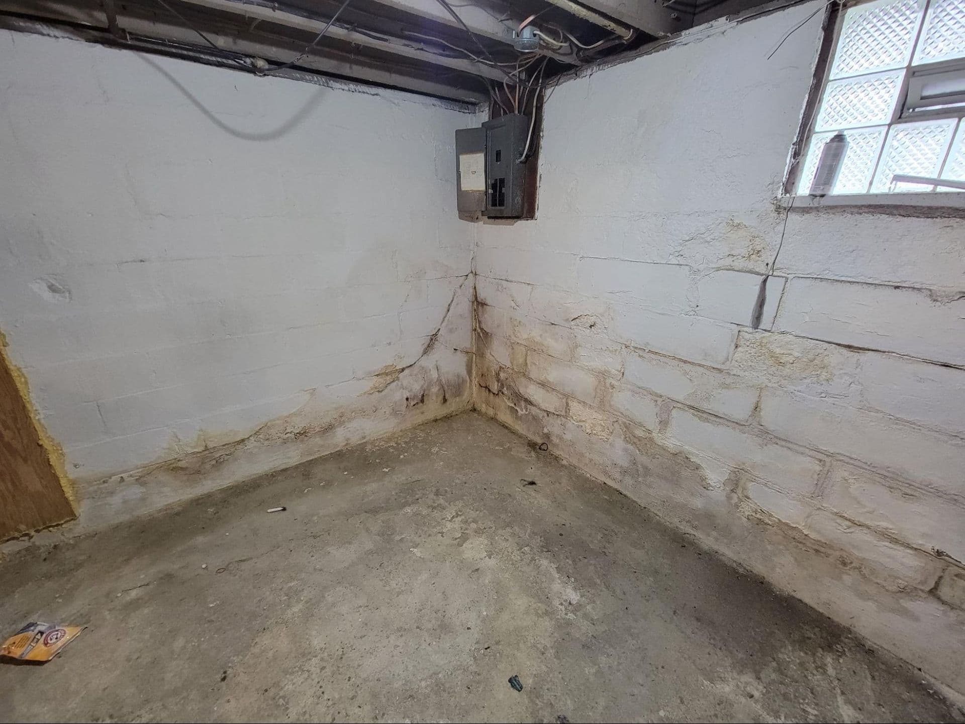 Basement corner with cracked and stained concrete foundation walls showing water damage and efflorescence, concrete floor, exposed electrical panel, and small window with security grate