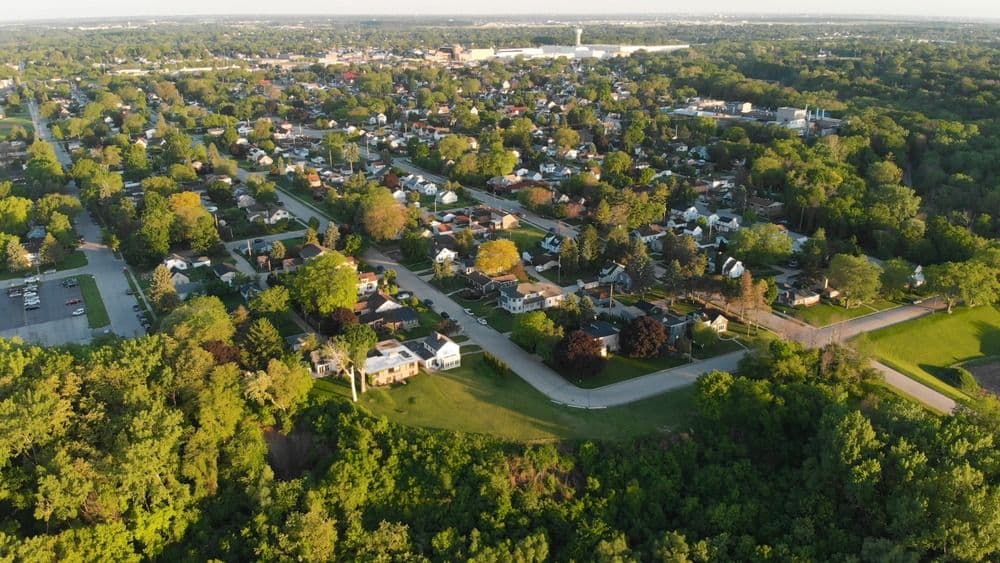 Aerial view of suburban neighborhood with single-family homes, tree-lined streets, green spaces, and commercial buildings in the distance