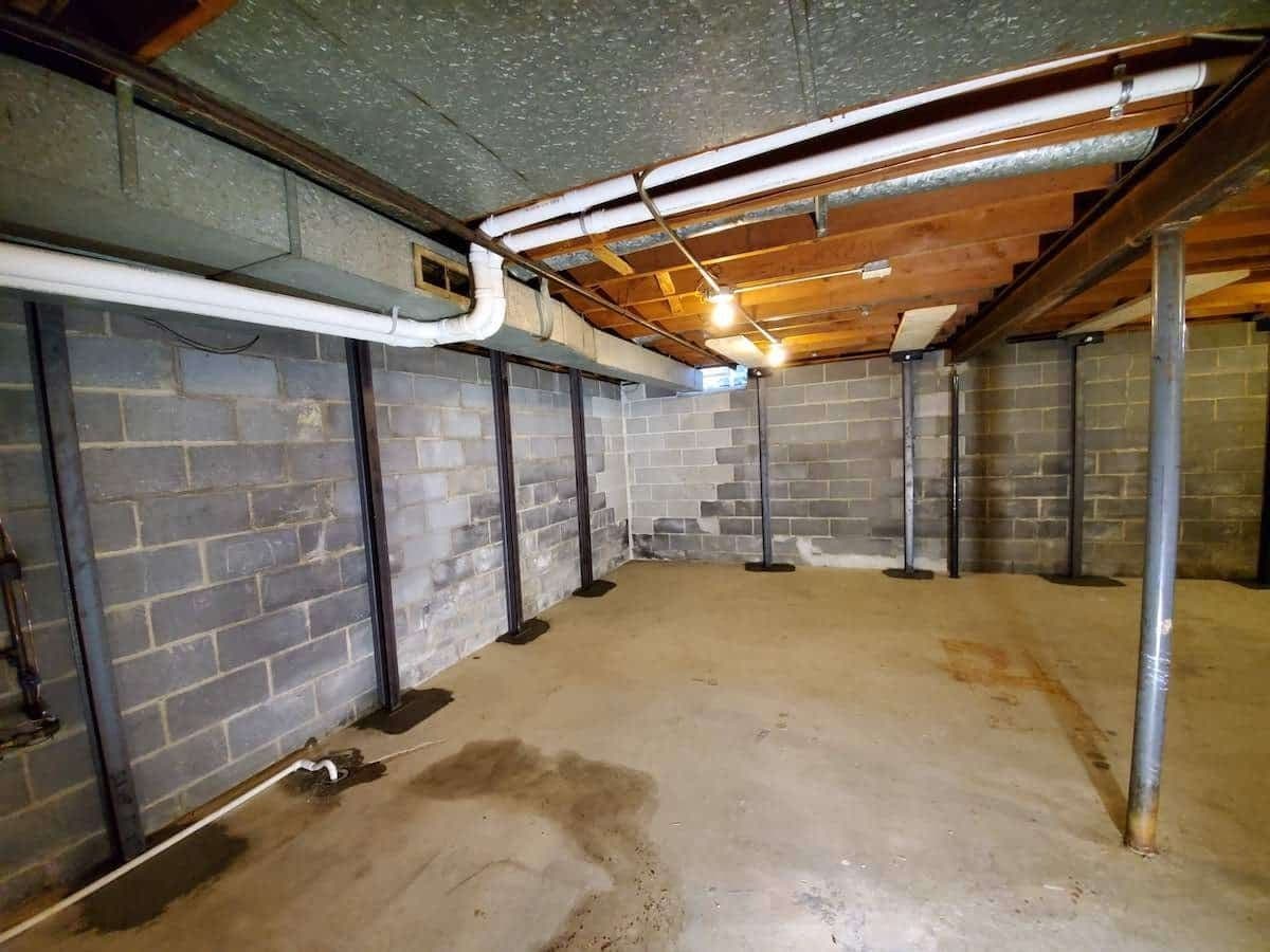 Empty basement room with concrete floor, brick walls, exposed wooden beams, fluorescent lighting, and metal support posts