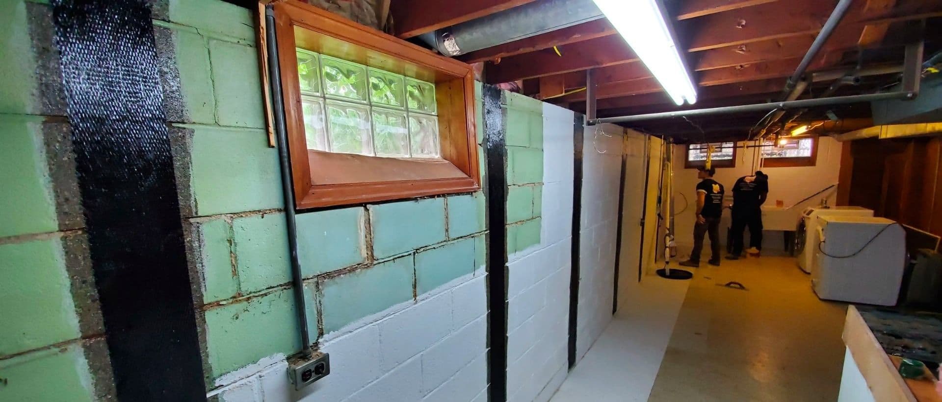 Interior of basement or underground space with green and white painted brick walls, wooden-framed window, exposed ceiling with pipes and fluorescent lighting, and two people standing in the distance