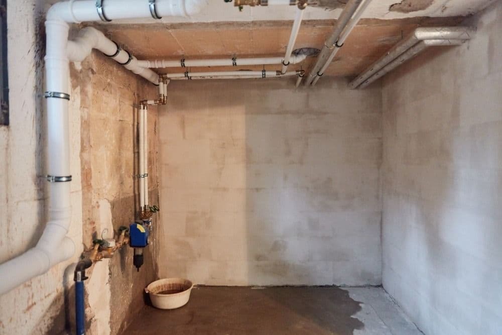 Unfinished basement room with exposed concrete walls and ceiling, white PVC plumbing pipes running overhead, and a plastic basin on the floor