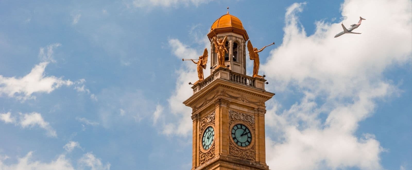 Golden clock tower with ornate dome and angel statues against blue sky with clouds, airplane flying overhead