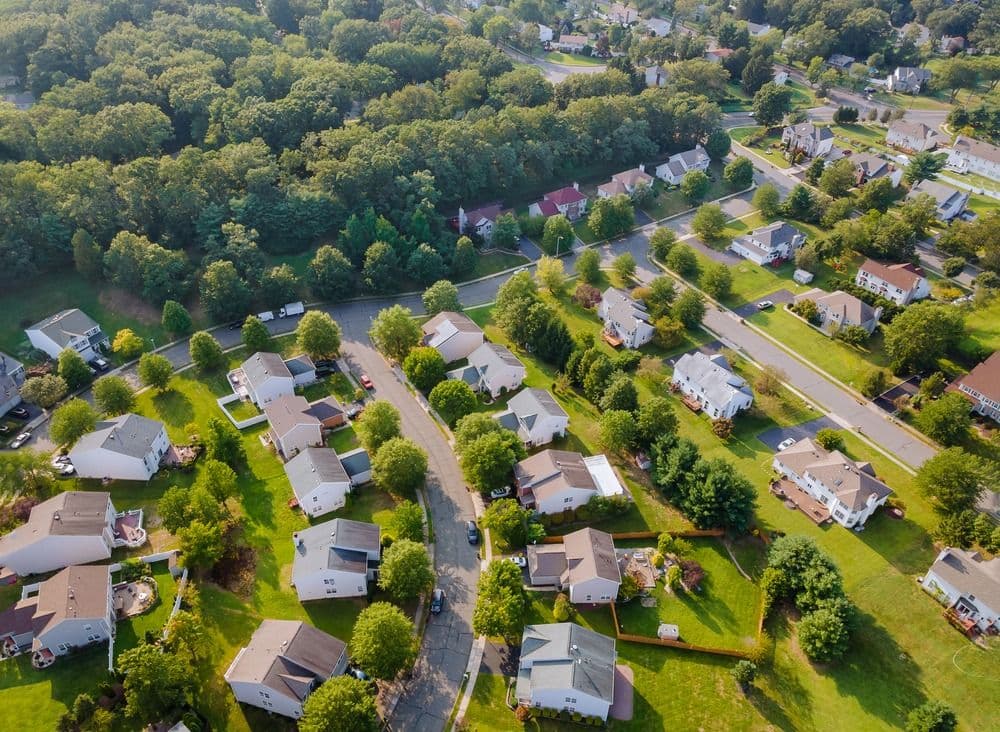 Aerial view of suburban residential neighborhood with single-family homes, curved streets, mature trees, and green lawns, bordered by dense forest