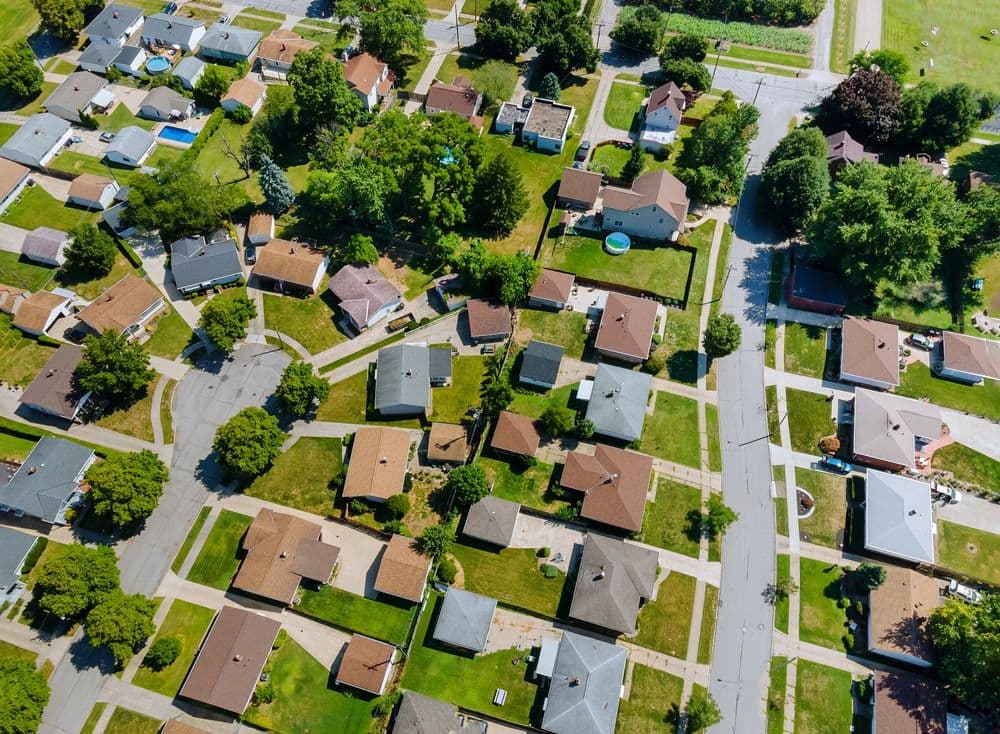 Aerial view of a residential suburban neighborhood with single-family homes, driveways, green lawns, and mature trees lining streets