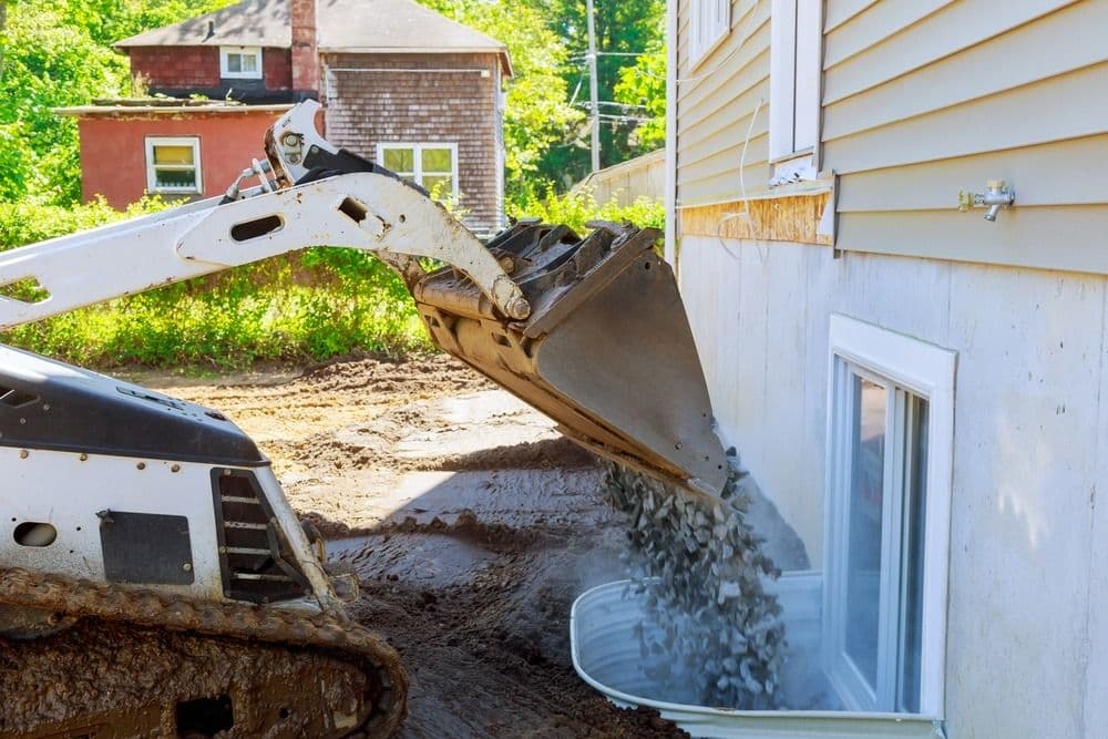 Excavator operator using bucket to dump gravel against residential foundation during site work near houses