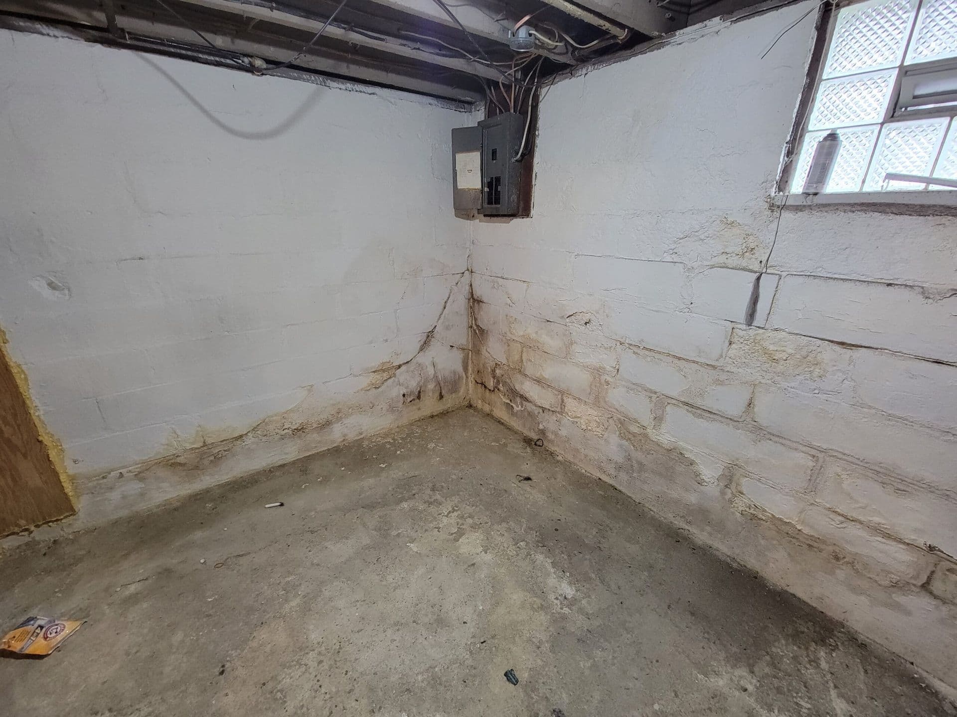 Basement corner with water stains and efflorescence on white painted concrete block walls, exposed ceiling beams, small window, and electrical outlet showing moisture damage