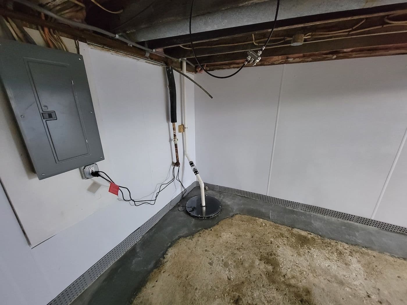 Basement crawl space with sump pump system, electrical panel on white wall, and exposed wooden joists overhead
