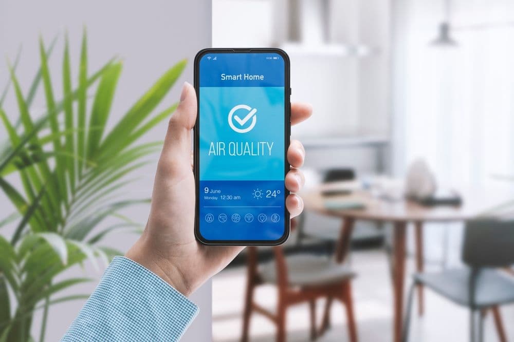 Hand holding smartphone displaying Smart Home app with Air Quality monitoring showing checkmark, date, temperature 24°C, and control icons in modern living room.