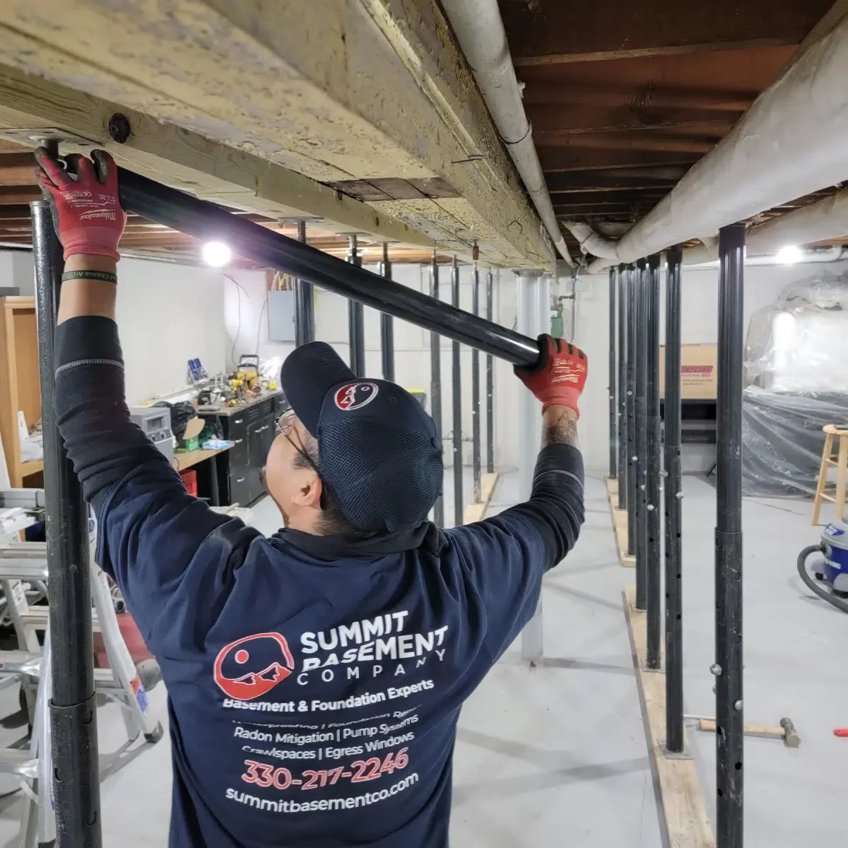 Worker from Summit Basement Company installing a support beam in an unfinished basement, wearing red work gloves and a navy shirt.