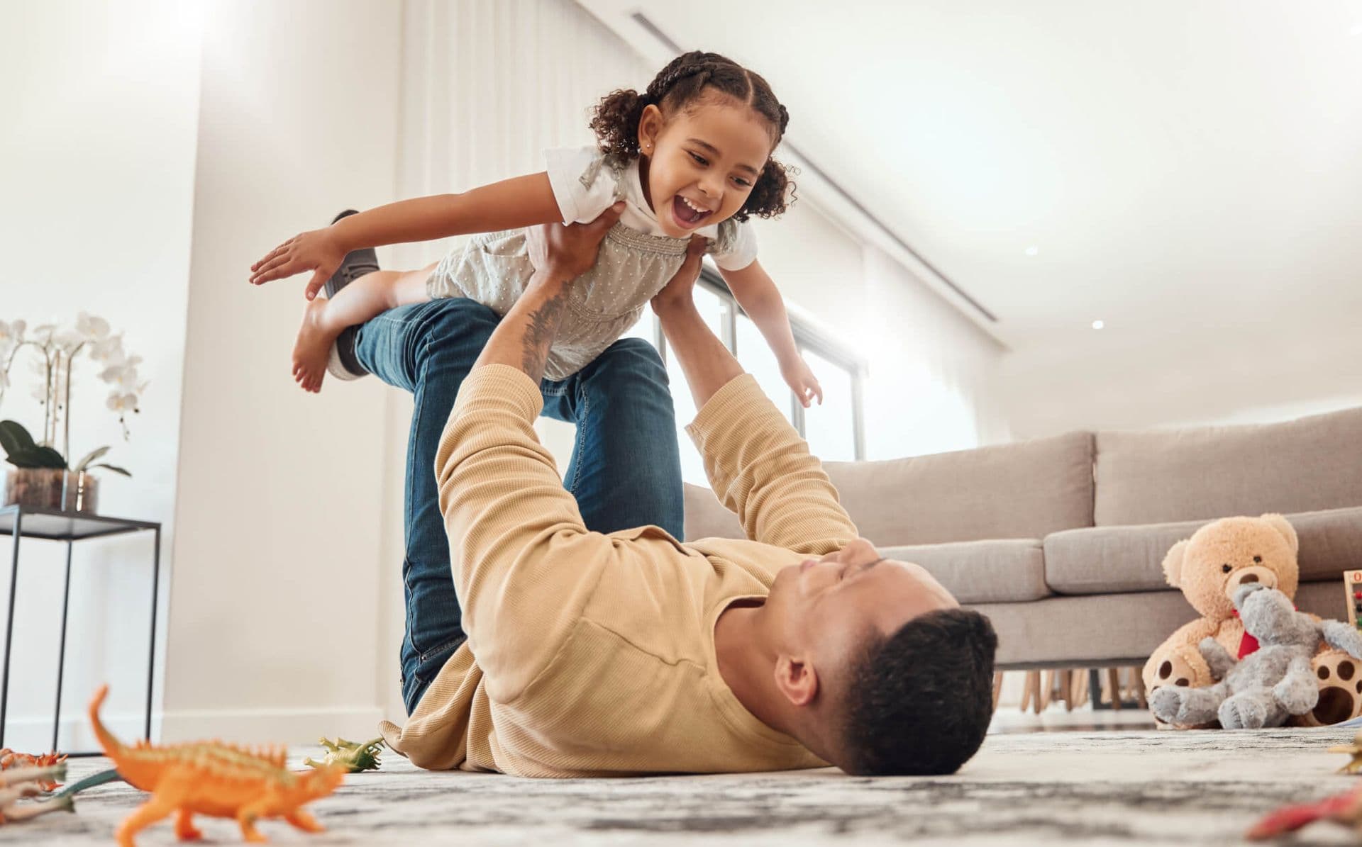 Father lying on floor lifting daughter in the air while playing together in a bright living room.