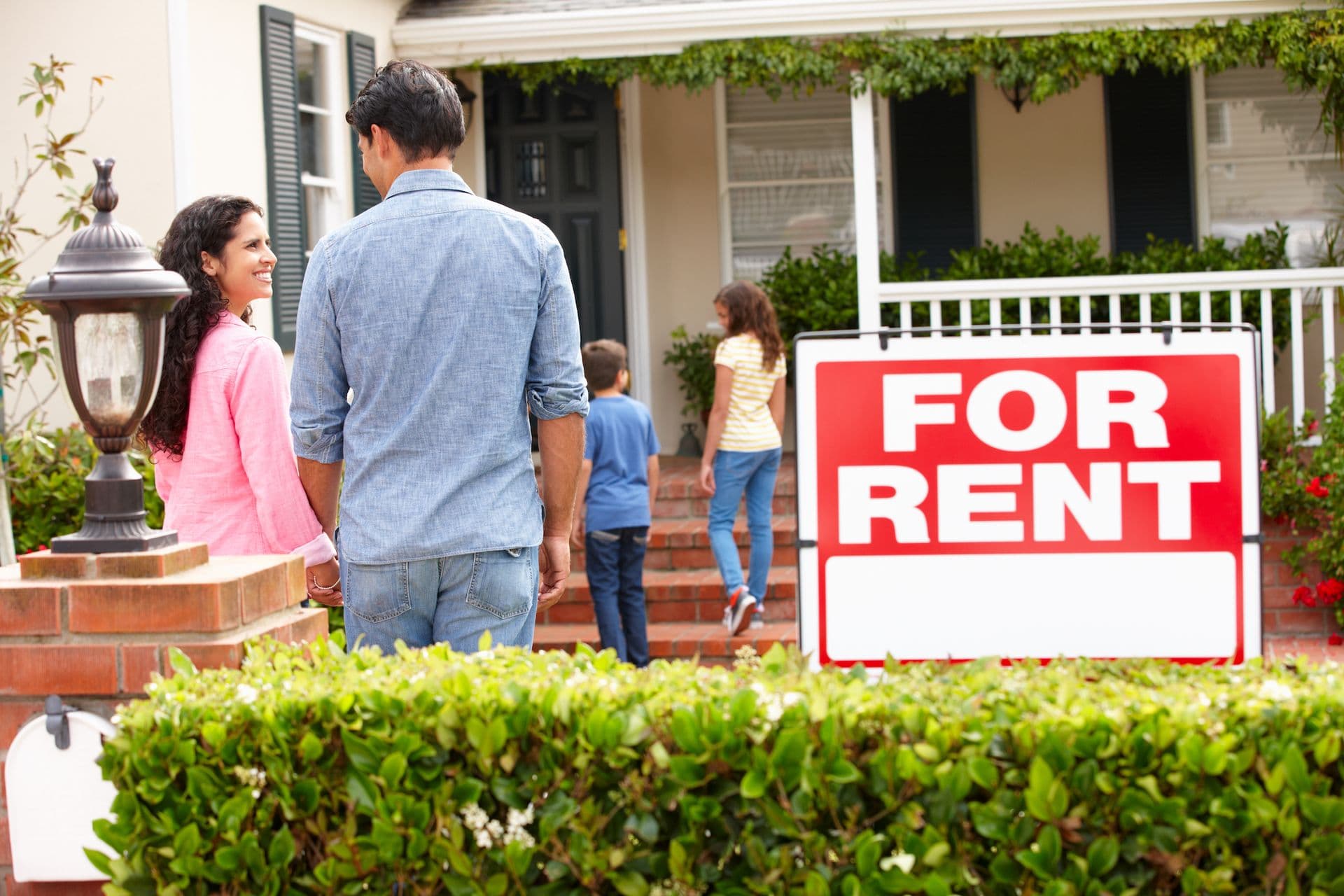 Family touring a house with a red "For Rent" sign displayed in the front yard.