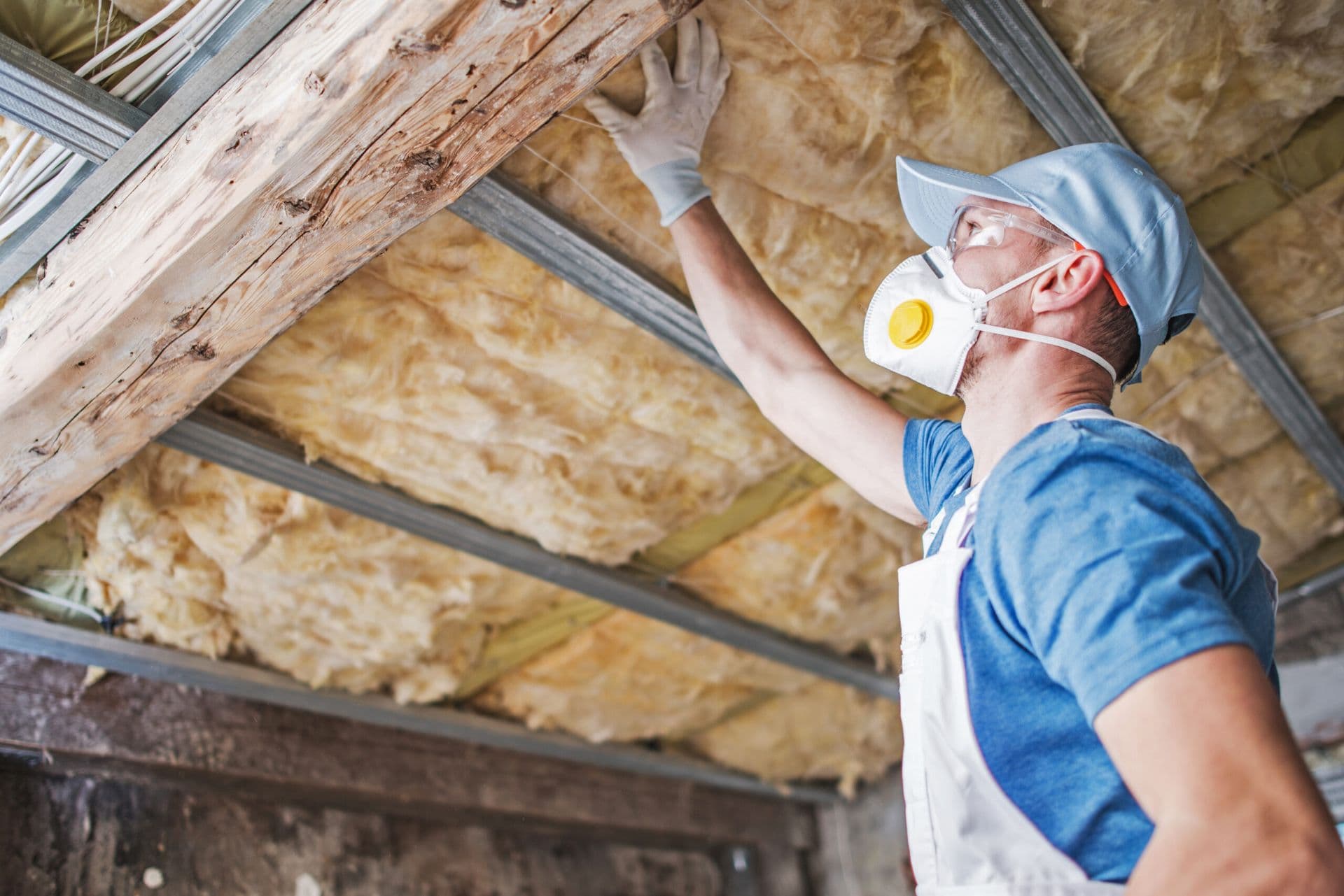 A qorker in hard hat and safety mask installing fiberglass insulation batts between ceiling joists and wooden beams.