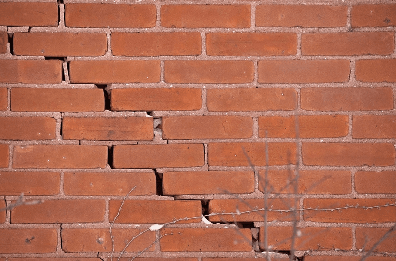 Cracked and damaged red brick wall showing structural issues with displaced and broken bricks along mortar lines.