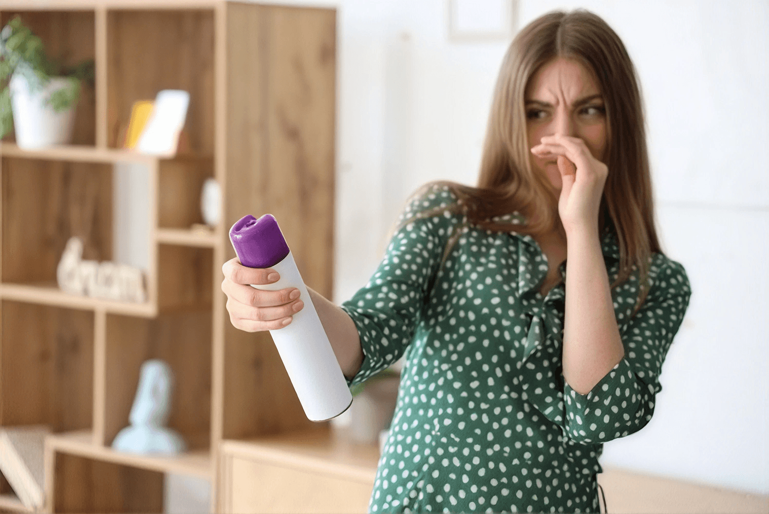 Woman in a green dress holding an air freshener spray with concerned expression, demonstrating unpleasant odor issue indoors.