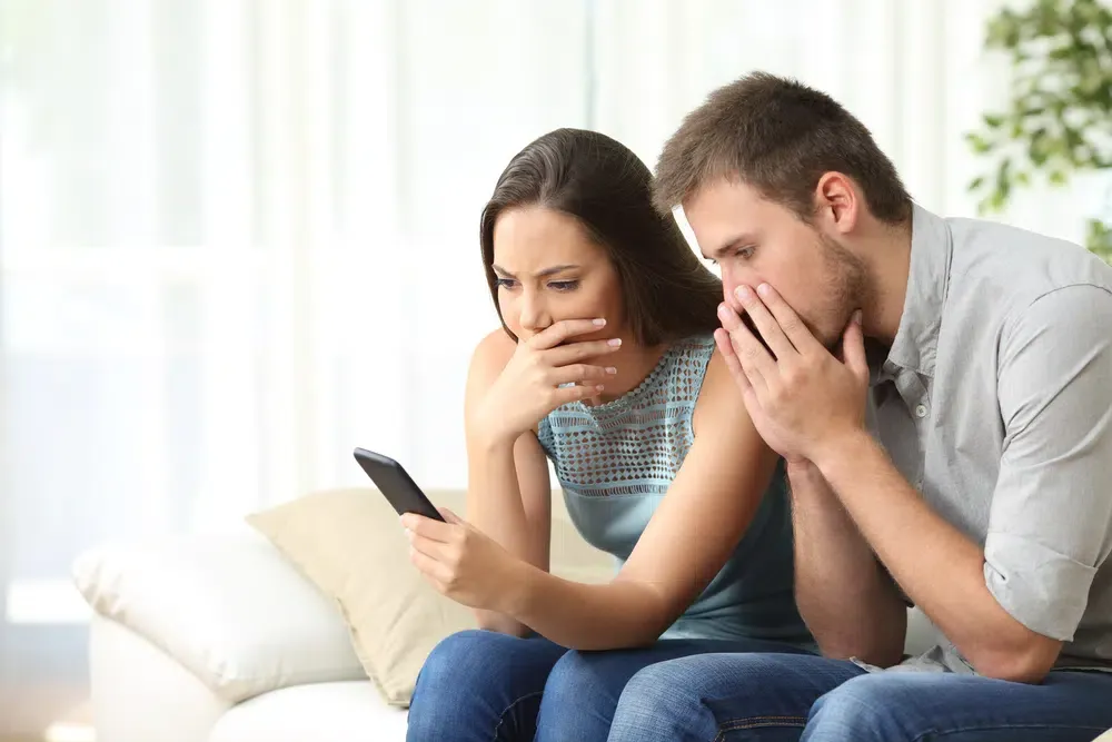 Concerned couple looking at smartphone together, both covering their mouths with worried expressions.