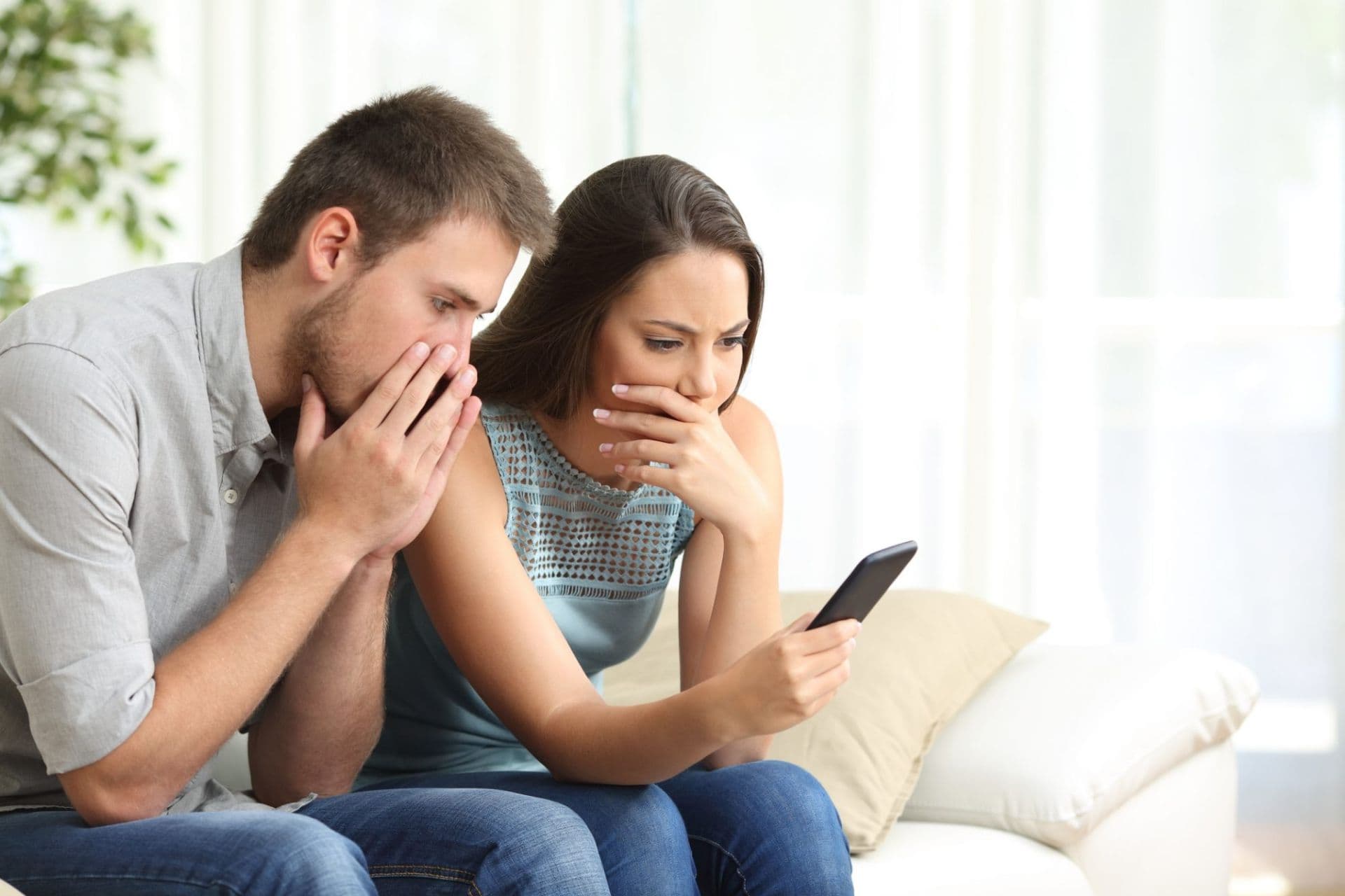 Concerned couple looking at smartphone together, both covering their mouths with worried expressions.