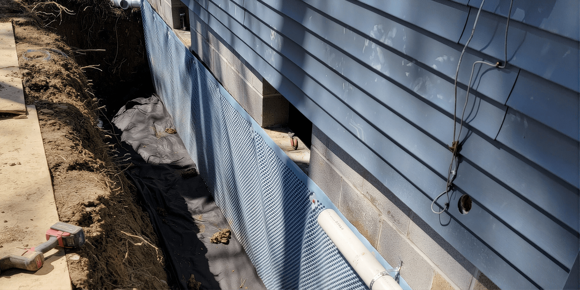 Newly-installed light gray reinforcement and waterproofing membrane installed around the foundation of a blue house.