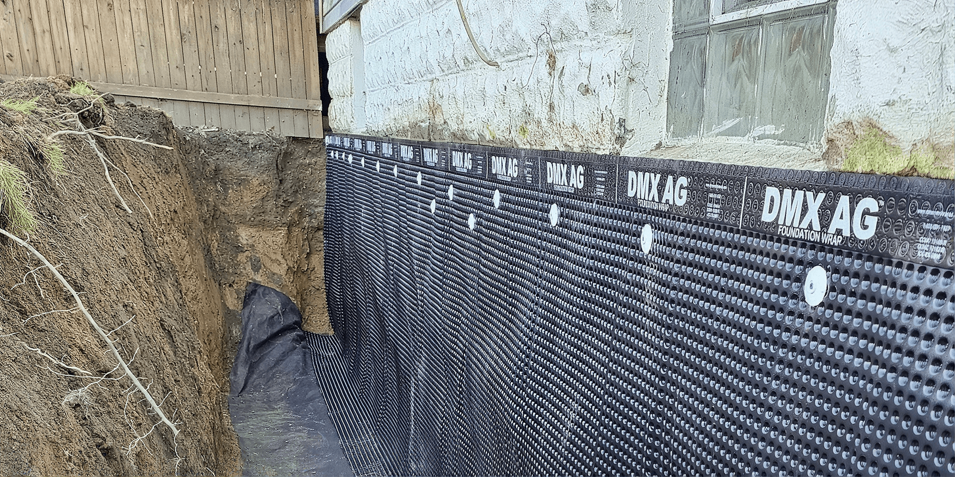 Black DMX AG foundation wrap membrane installed along excavated trench next to aged building foundation.
