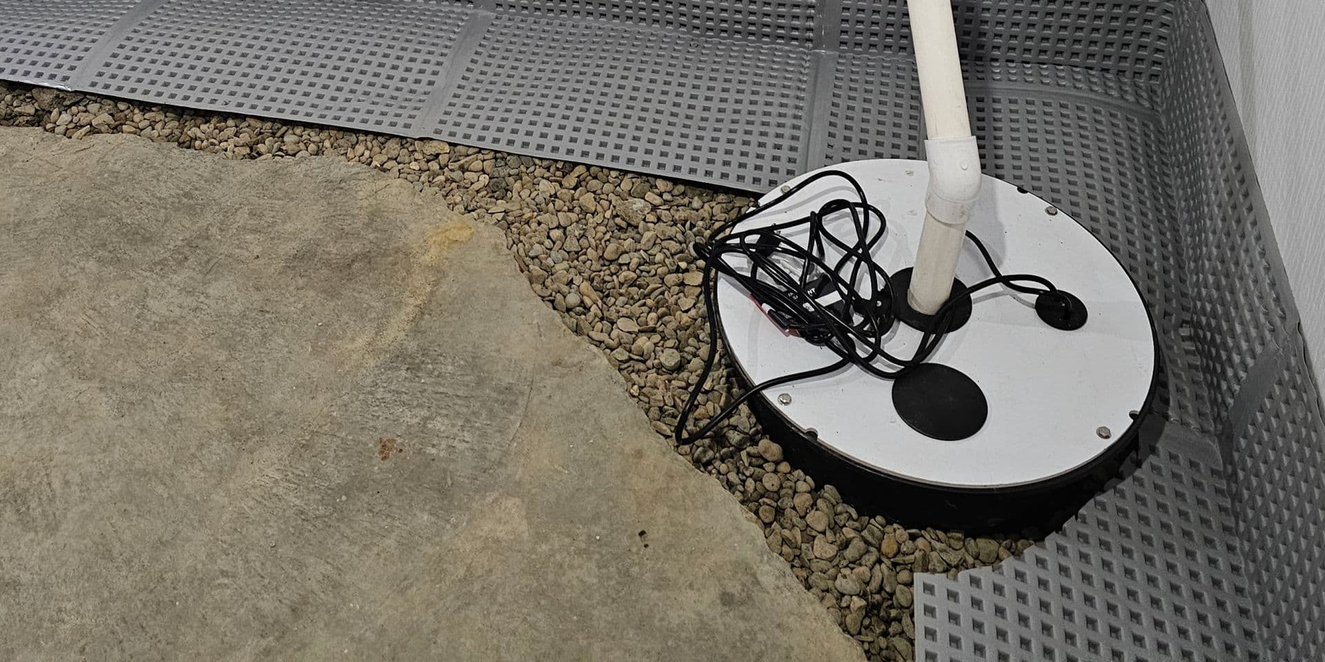 A sump pump system beside gravel and concrete surface with gray reinforcement material lining the edges along the wall.