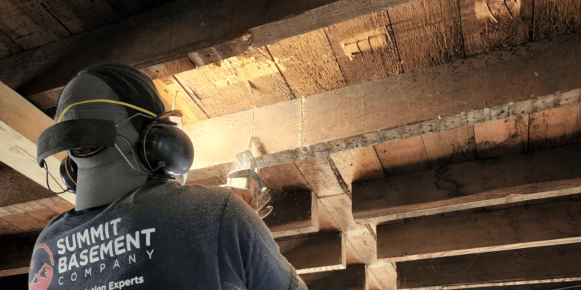 Worker from Summit Basement Company wearing safety gear works on wooden basement joists and beams.