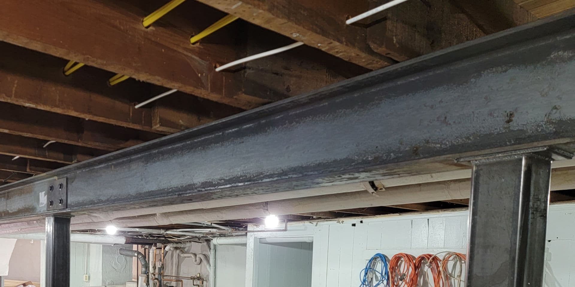 Exposed basement ceiling with steel I-beams and wooden joists, yellow and white safety markings on beams, LED lighting, and coiled electrical cords hanging on white wall below.