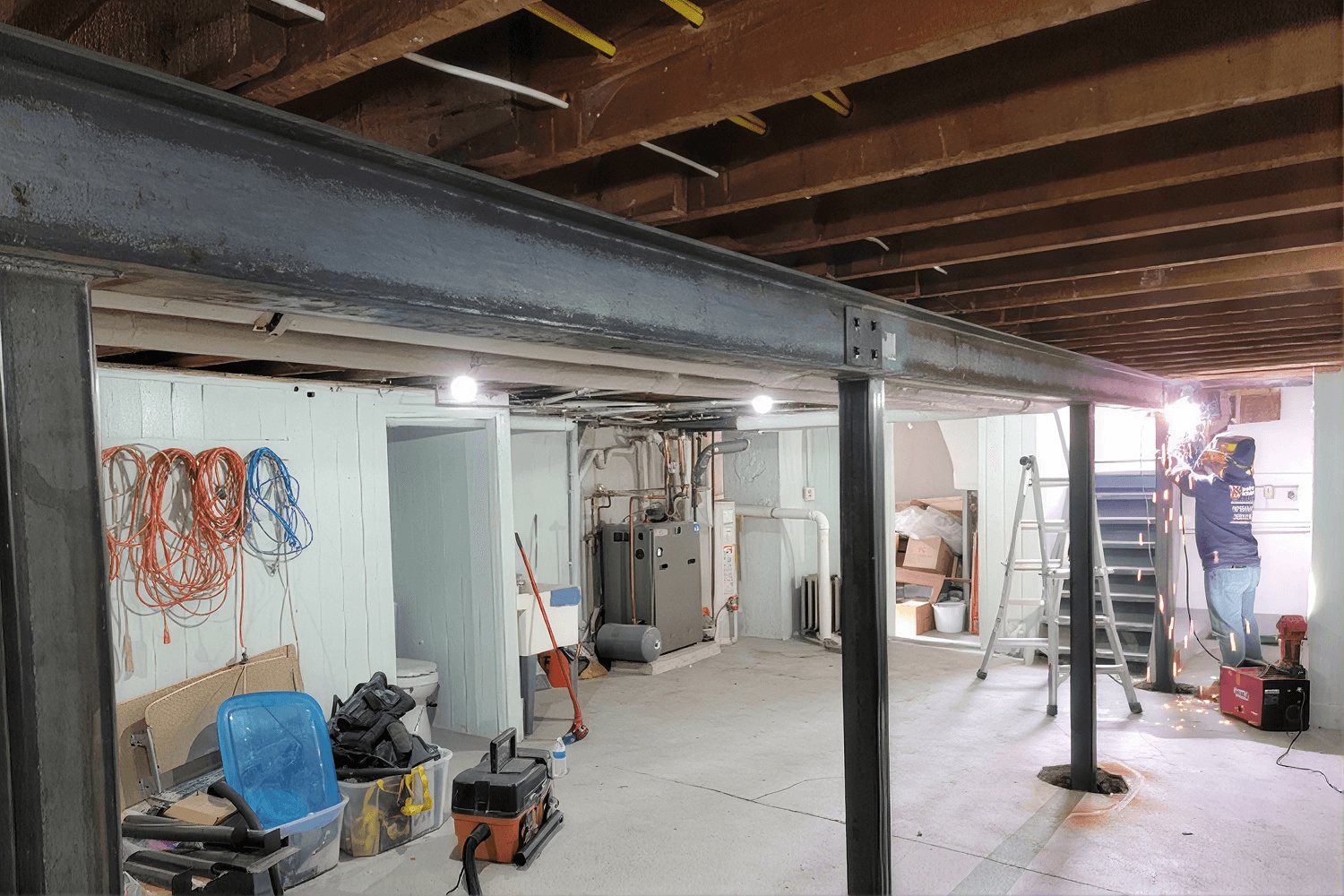 Unfinished basement under renovation with exposed wooden beams, construction materials, coiled cables, ladders, and utility equipment scattered throughout the space.