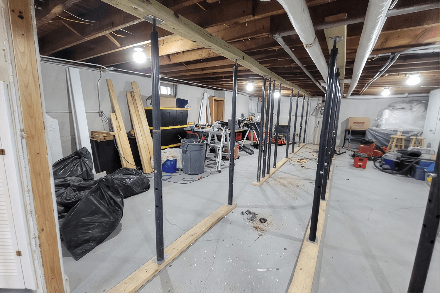 Unfinished basement workspace with metal support posts, wooden materials stacked along walls, black trash bags, tool storage containers, and various equipment scattered throughout.