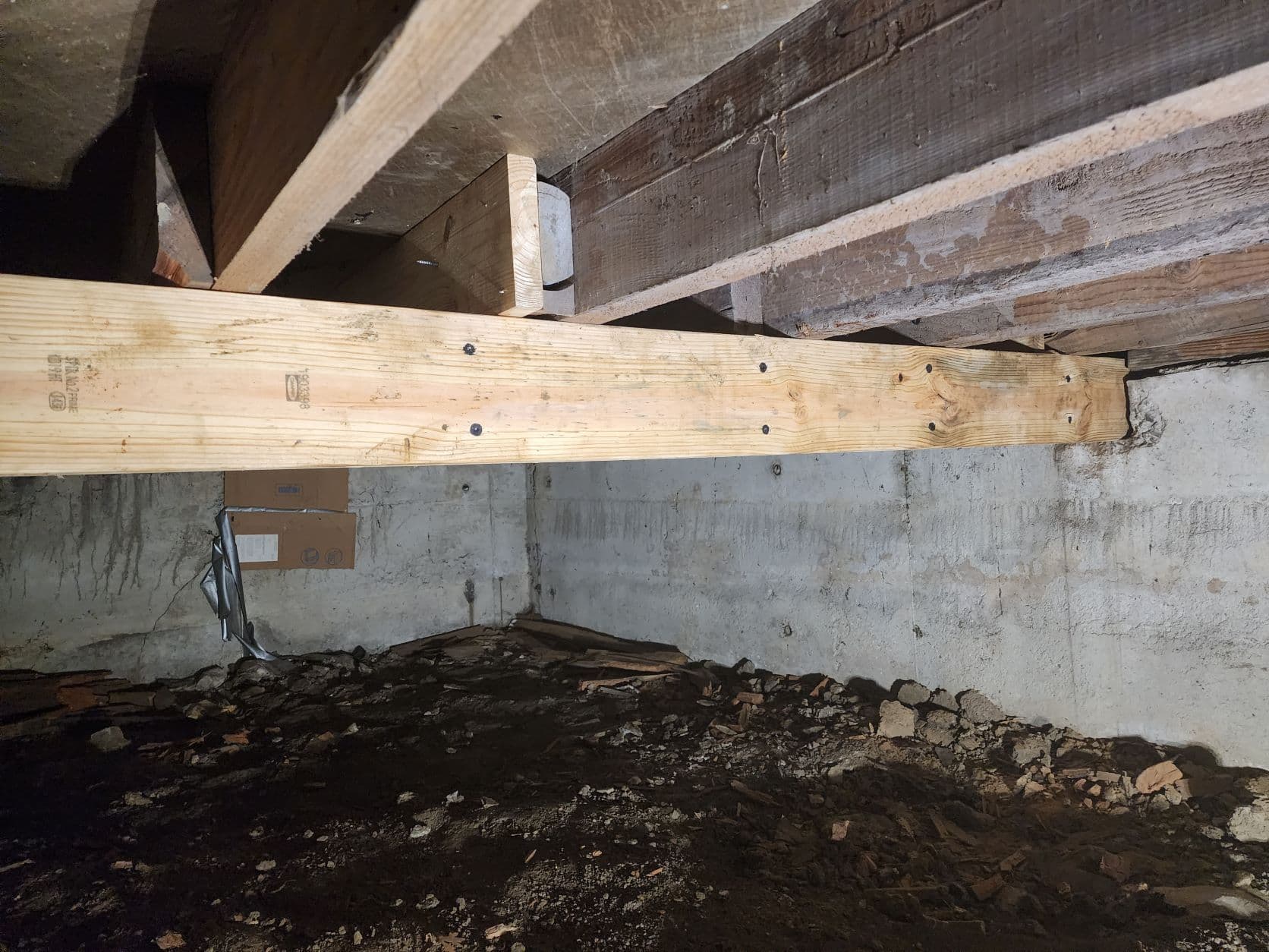 New wooden support beam installed in basement crawlspace with concrete foundation and exposed floor joists above.