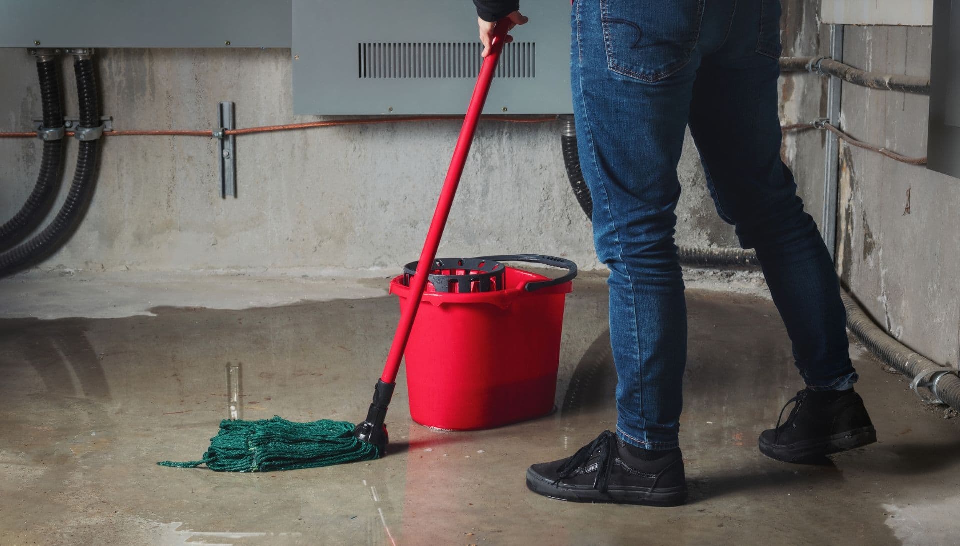 A person using a mop and red bucket to clean a concrete basement floor with piping and wiring visible on walls.